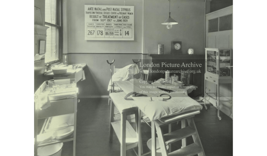 photograph from the London Archives, linked to their site, depicting the operating theatre of the Thavies Inn Infirmary, a chart on the wall reads 'Ante-natal and post-natal syphillis' and 'result of treatment of cases' with figures.